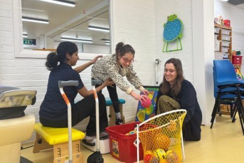 Girl reaching to put ball into a basket while standing on galileo vibration plate. With a physiotherapist to her left, and therapy assistant to her right, during a session at CPotential.